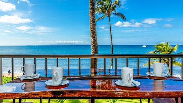 A table with four white cups and saucers on a wooden table overlooking a beach with palm trees and the ocean.
