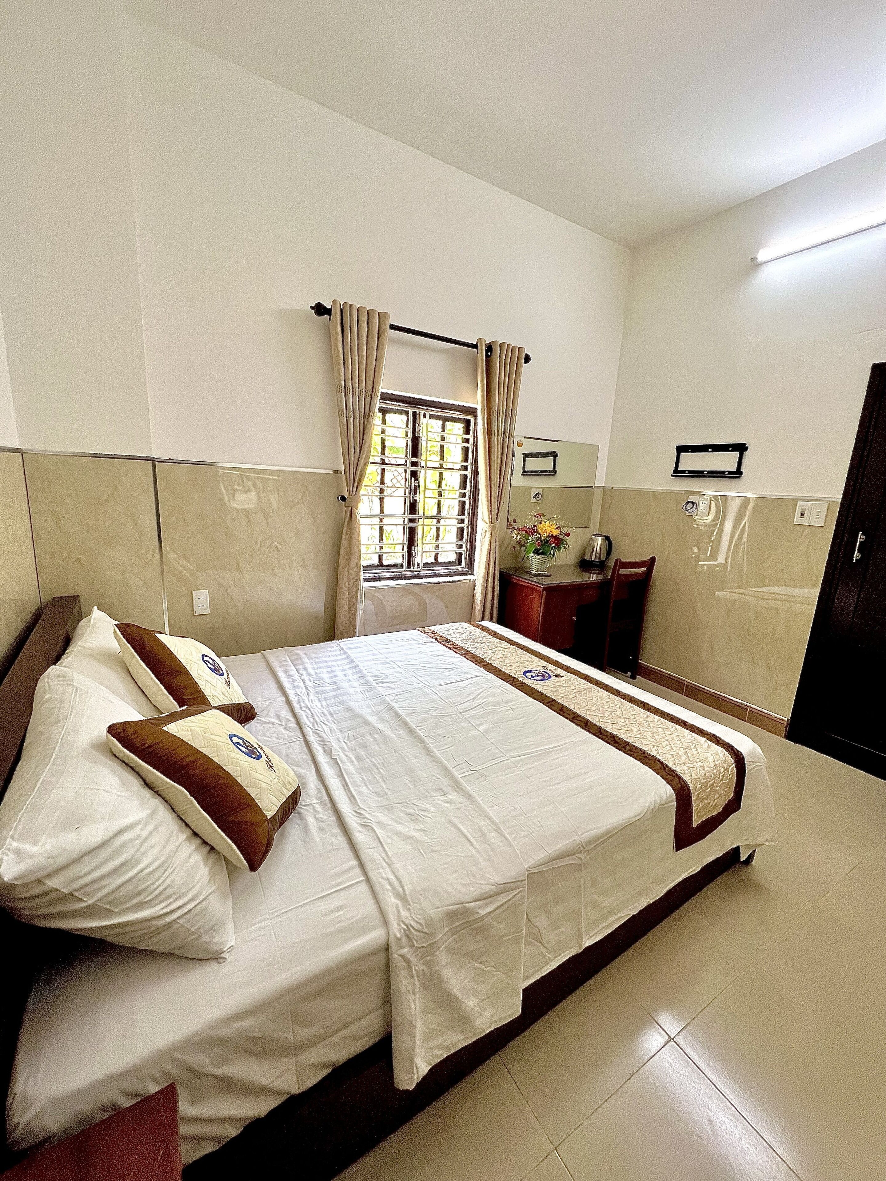 A hotel room with a bed, a window with curtains, a bedside table with a lamp, and a wall-mounted shelf.