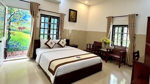 A spacious bedroom with a large bed, a desk, and a chair. There is a window with curtains, a painting on the wall, and an air conditioning unit. - Ngo Family Apartment (Da Nang)
