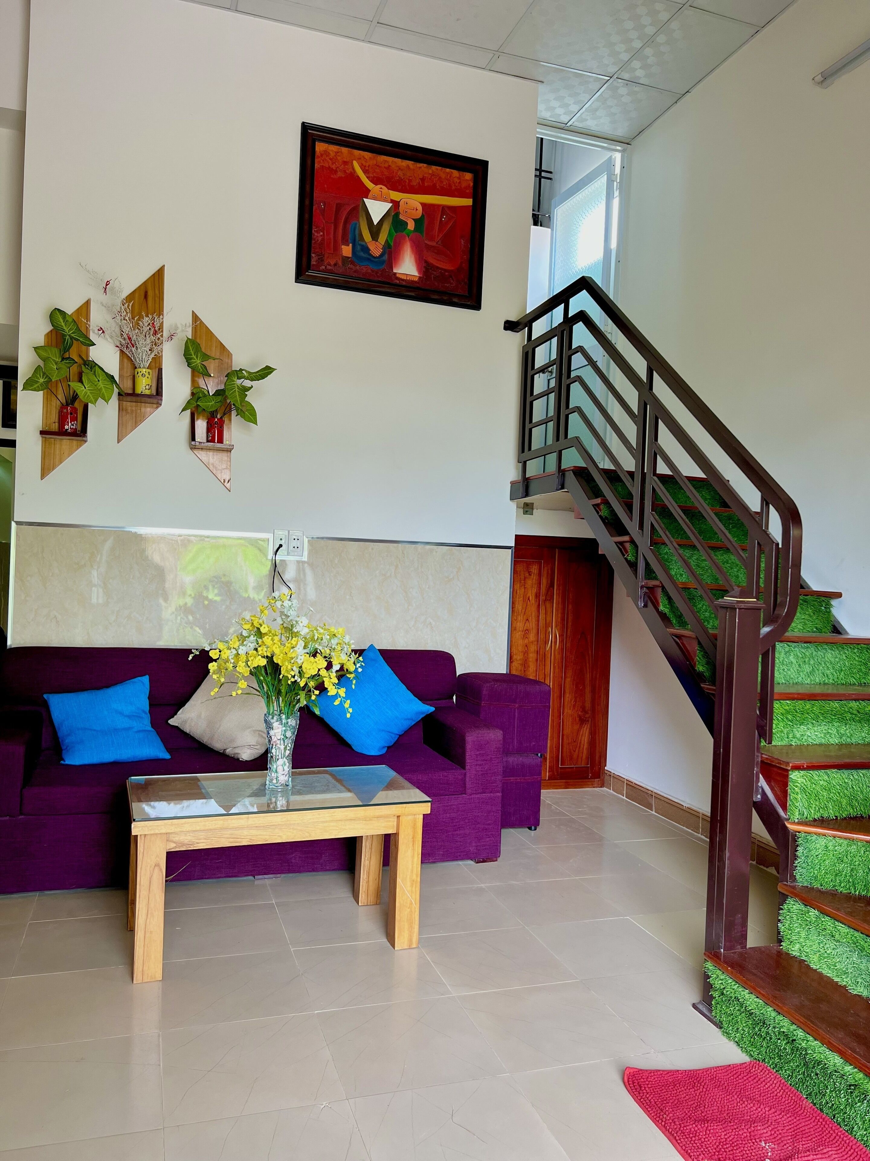 Un salón con un sofá morado, una mesa de centro de madera y una escalera con peldaños cubiertos de césped artificial verde.