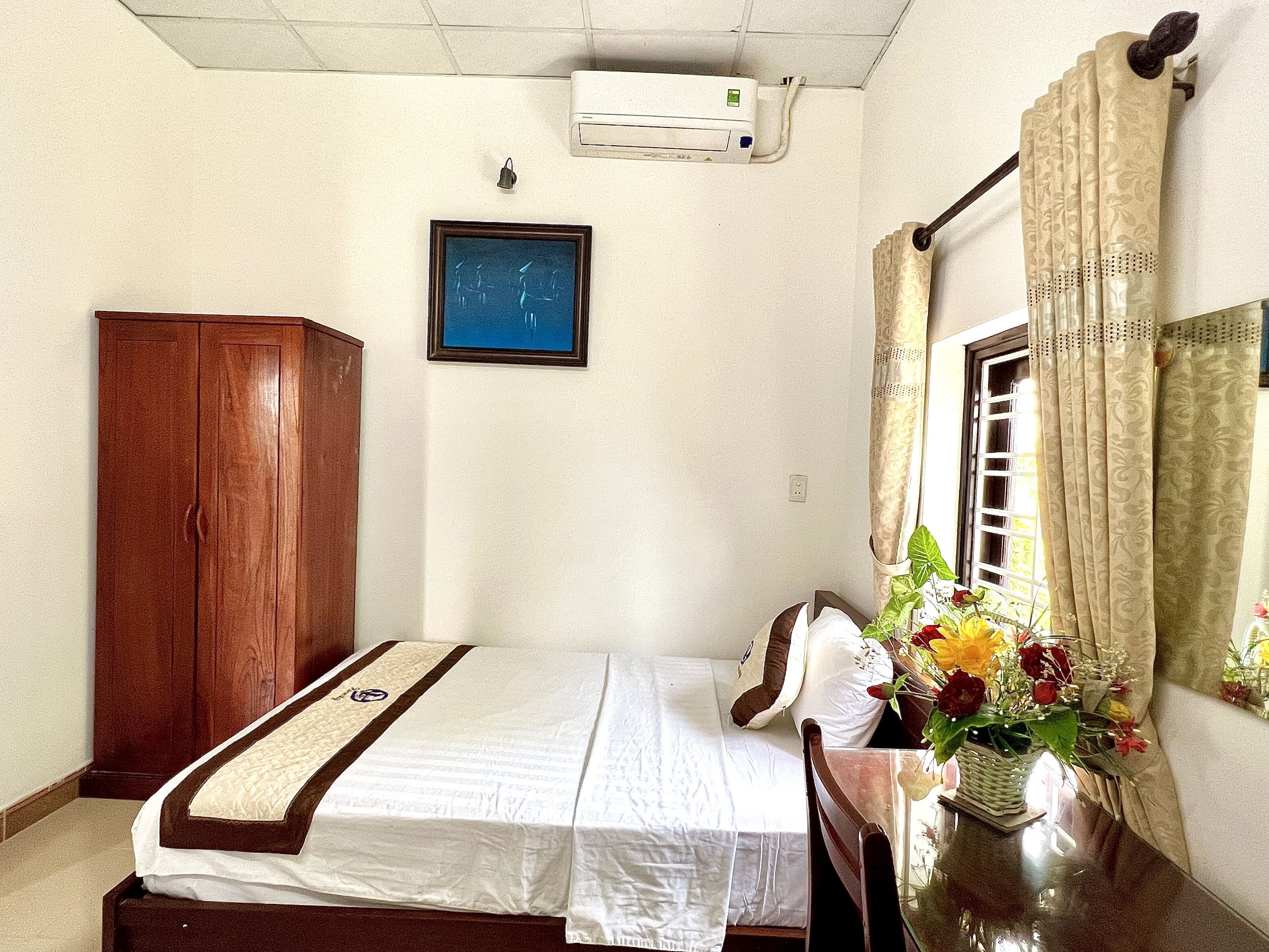 A bedroom with a bed, a wooden wardrobe, a wall-mounted air conditioner, a framed picture, and a window with curtains.