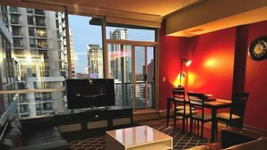 A modern living room with a leather sofa, a TV, a dining area with chairs, and a large window offering a city view. - 2bedroom/2washroom condo close to everything + Free Parking (Calgary)
