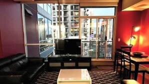 A living room with a black leather sofa, a coffee table, and a TV. There is a large window with a view of buildings. - 2bedroom/2washroom condo close to everything + Free Parking (Calgary)