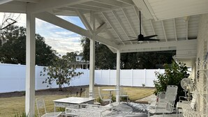Une terrasse couverte, dotée de meubles en métal blanc, d’un ventilateur de plafond et offrant une vue sur une clôture blanche et des arbres en arrière-plan.