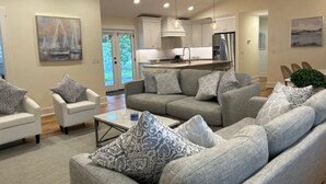 Un salon moderne avec un canapé gris, une table basse et des coussins à motifs. On y trouve également un coin cuisine avec des meubles blancs et des appareils électroménagers en acier inoxydable.