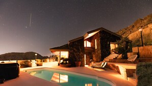 Vista nocturna de una casa con piscina, fachada de piedra y zona de estar al aire libre.