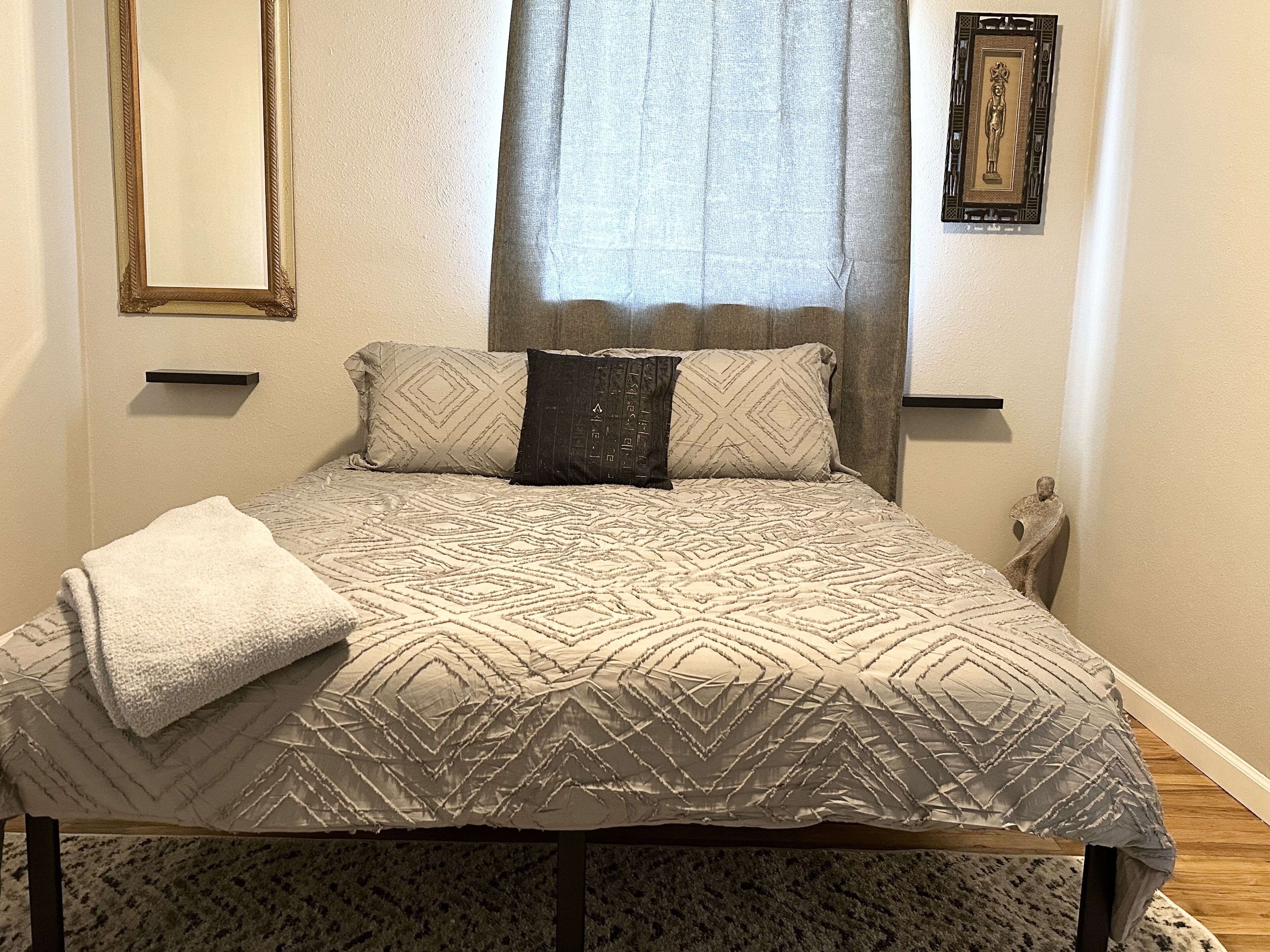 Un llit amb un estriat, dos coixins, una tovallola doblegada, una tauleta de nit, una fotografia emmarcada a la paret i una finestra amb cortines.