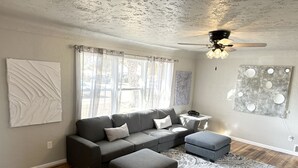 Ein Wohnzimmer mit Ecksofa, Ottoman, gemustertem Teppich, Deckenventilator und Kunstwerken an den Wänden.