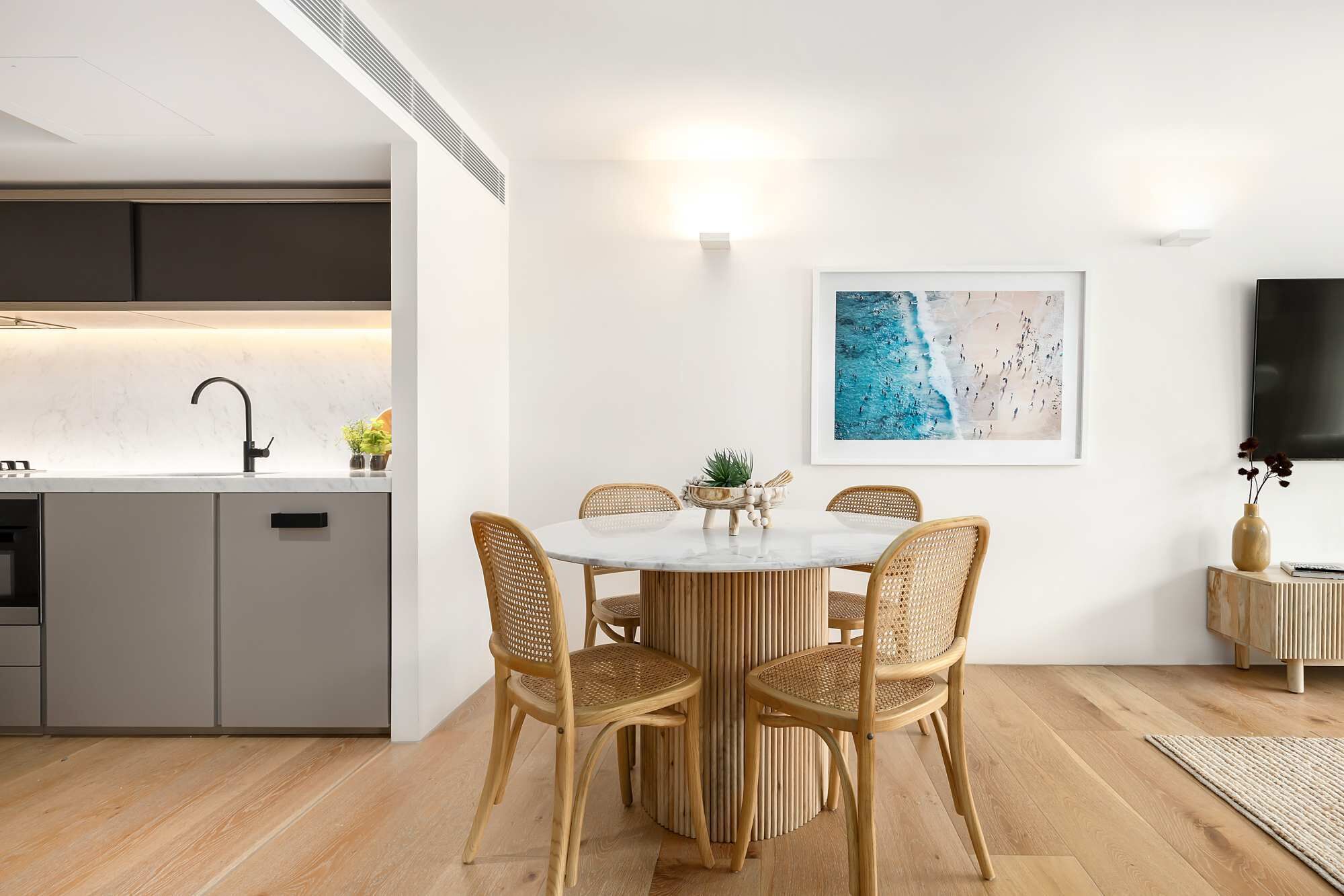 Une cuisine moderne dotée d’un four encastré, un coin repas avec une table ronde et des chaises en osier, un tableau accroché au mur et un téléviseur à écran plat.
