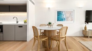 A modern kitchen with a built-in oven, a dining area with a round table and wicker chairs, a painting on the wall, and a flat-screen TV. - Bondi Breeze (Bondi)