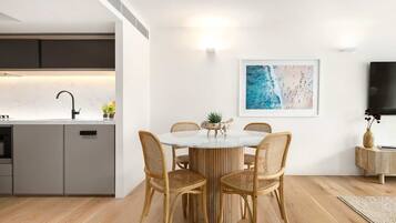 Une cuisine moderne dotée d’un four encastré, un coin repas avec une table ronde et des chaises en osier, un tableau accroché au mur et un téléviseur à écran plat.