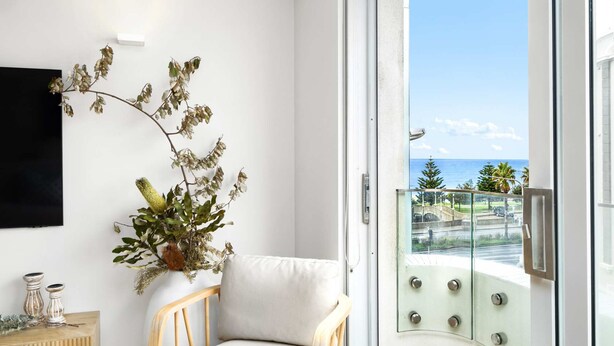 A modern interior with a white wall, a wooden chair, a white sofa, a potted plant, and a balcony with a view of the sea. - Bondi Breeze (Bondi)