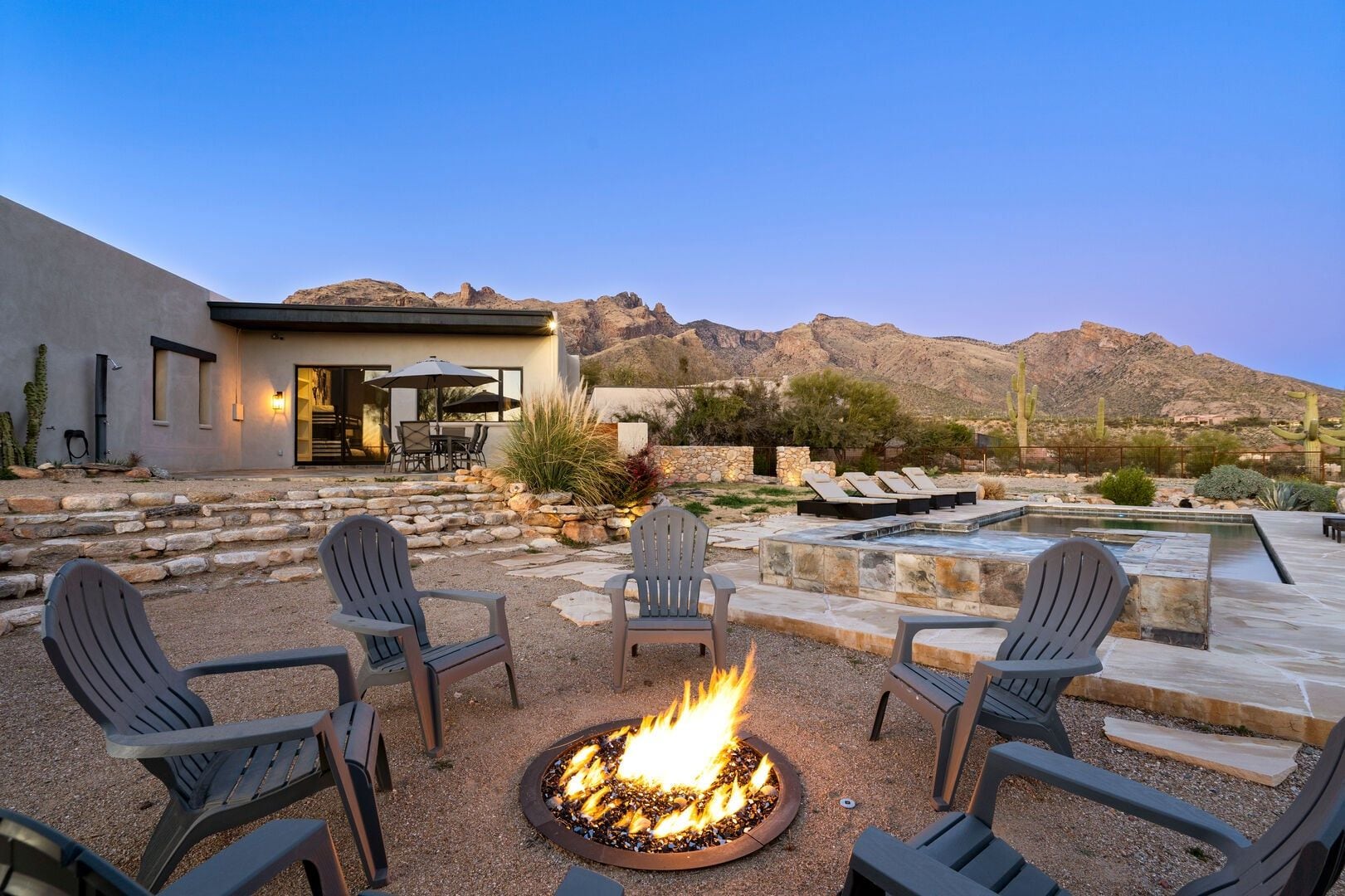 A modern outdoor patio with a fire pit, seating, and a pool area, surrounded by desert landscape and mountains.