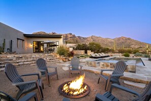 Un patio moderno al aire libre con una chimenea, asientos y una zona de piscina, rodeado por un paisaje desértico y montañas.