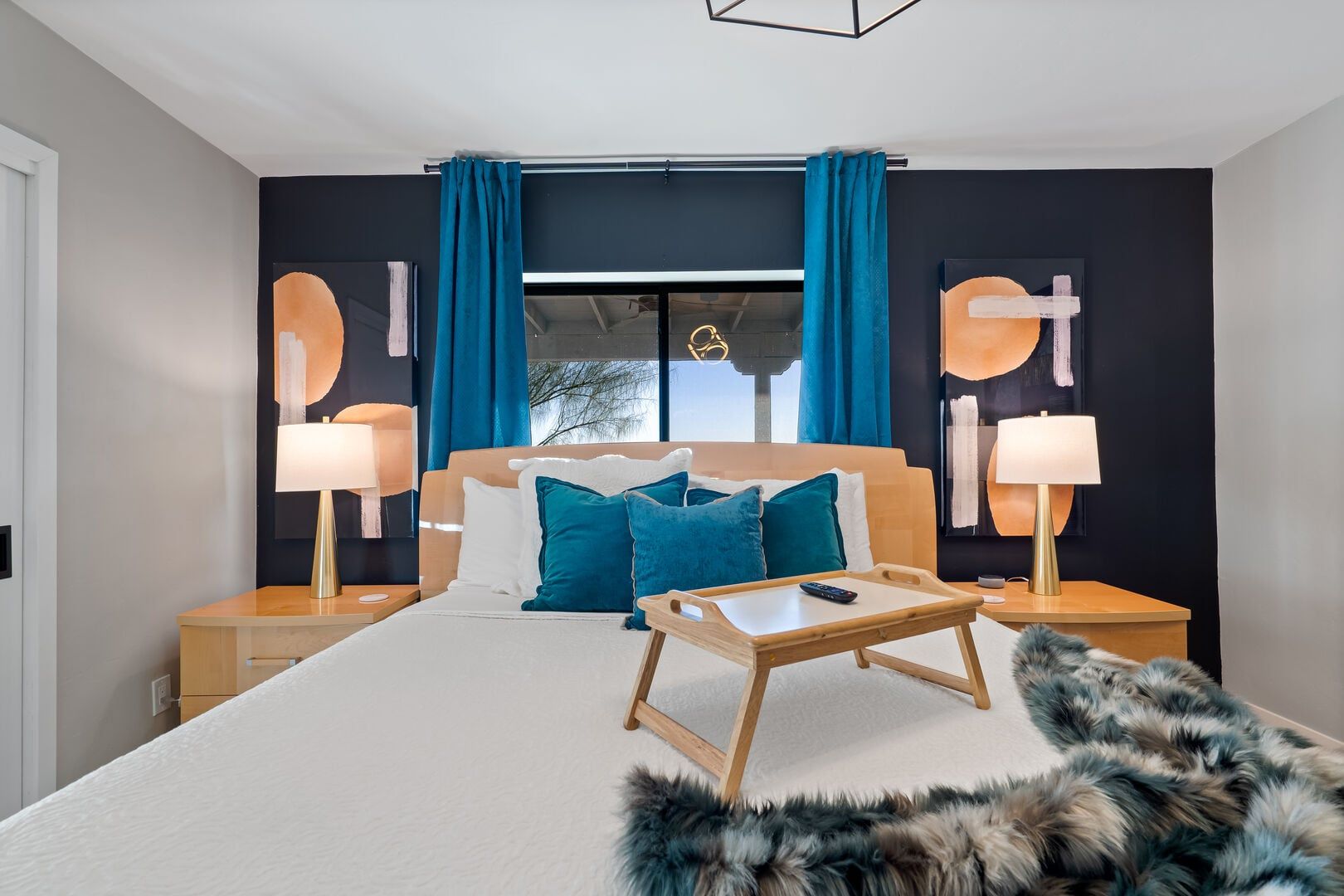 Une chambre d’hôtel moderne avec un grand lit, deux tables de chevet avec des lampes, une petite table en bois et un plaid en fourrure sur le sol.