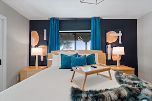 Una habitación de hotel moderna con una cama grande, dos mesitas de noche con lámparas, una pequeña mesa de madera y un mantón de piel en el suelo.