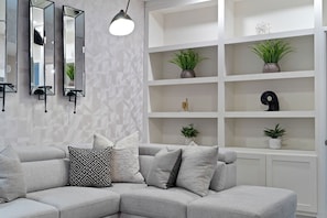 A modern living room with a grey sectional sofa, multiple cushions, a geometric patterned pillow, a wall-mounted mirror, a pendant light, and a white shelving unit with plants and decorative items. - Catalina Clubhouse | Pool, Spa, Pickleball & Basketball Court (Tucson)