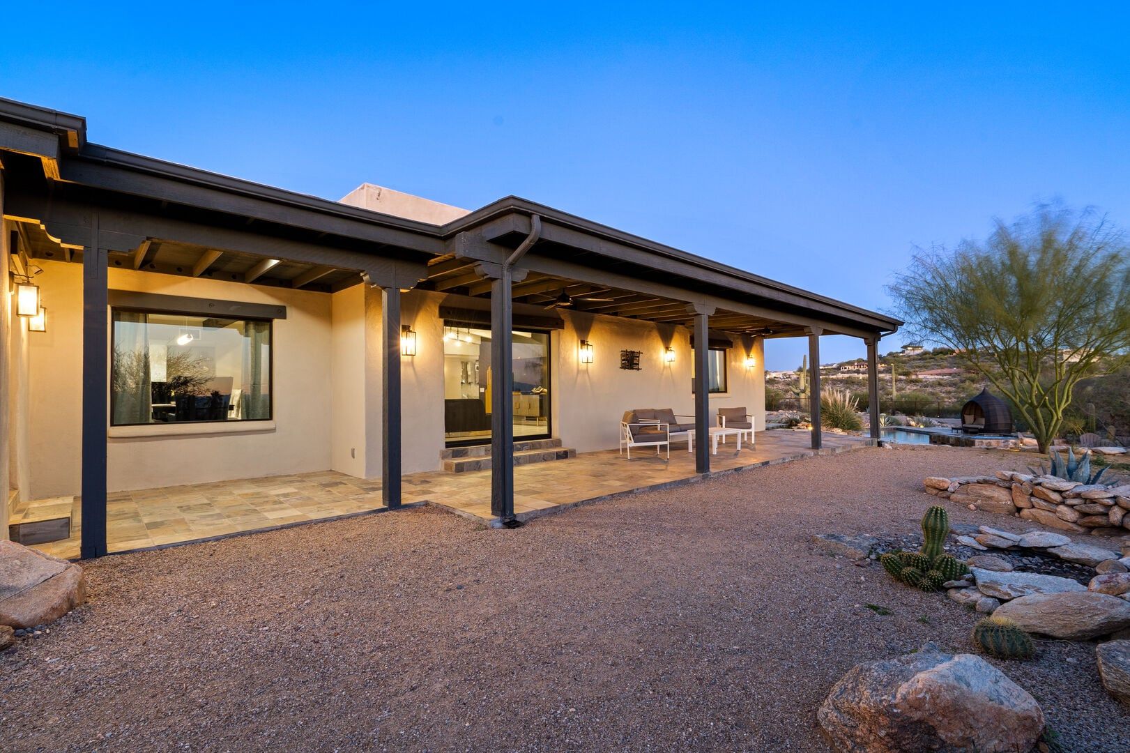 A modern house with a covered patio, outdoor seating, and a pool in the background.
