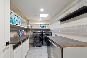 A compact laundry room with two front-loading washing machines, a drying rack, and storage shelves.