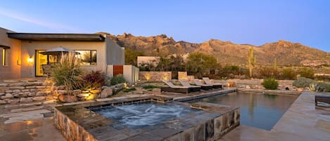 A modern outdoor pool area with a hot tub, lounge chairs, and a stone pathway leading to a building with a glass door.
