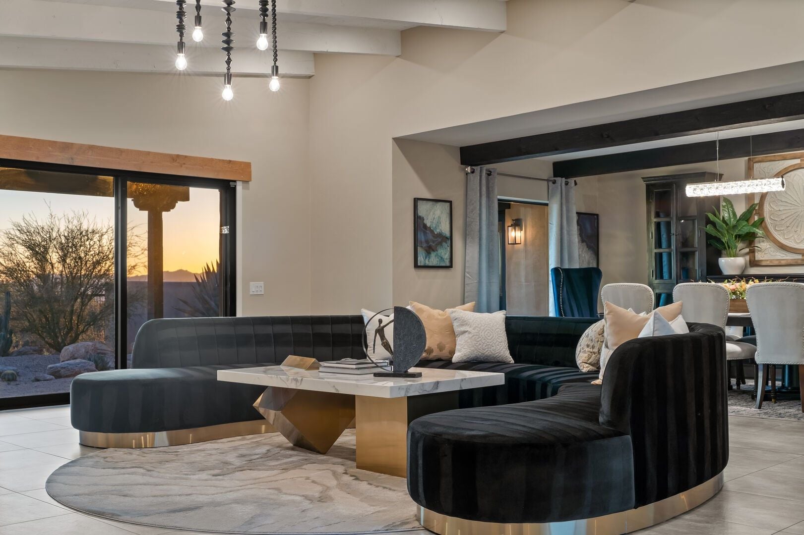 A modern living room with a large window, a black sofa set, a marble coffee table, and a dining area with a chandelier.