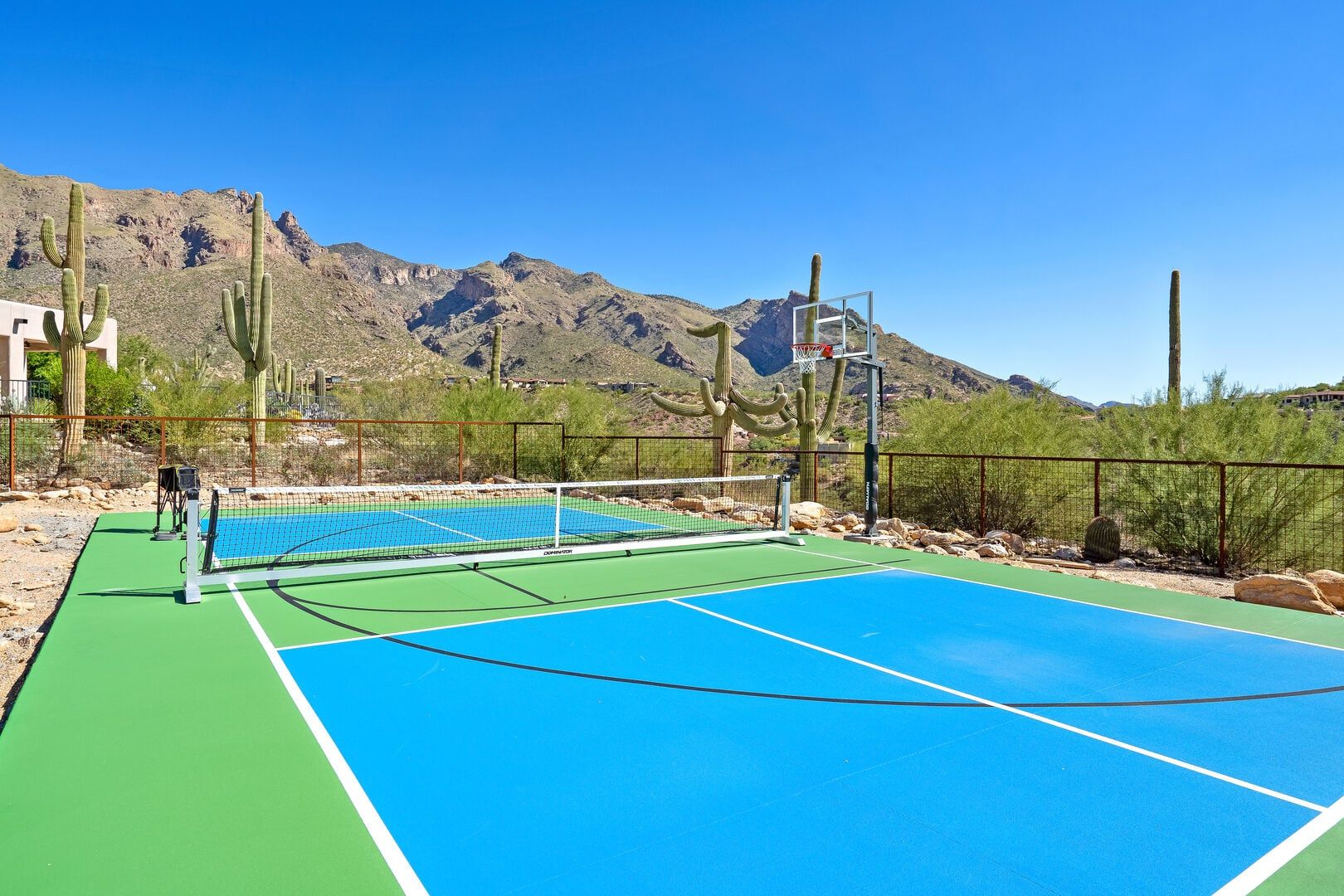 An outdoor tennis court with a net, surrounded by a fence, and cacti in the background.