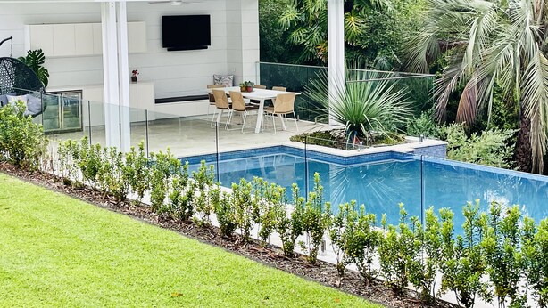 A modern outdoor patio with a pool, surrounded by lush greenery and a white building with a flat roof.