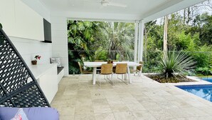 A modern outdoor patio with a dining table and chairs, surrounded by lush greenery and a swimming pool.