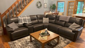 A living room with a leather sectional sofa, a wooden coffee table, a patterned rug, a staircase, and a dining area with wooden chairs.