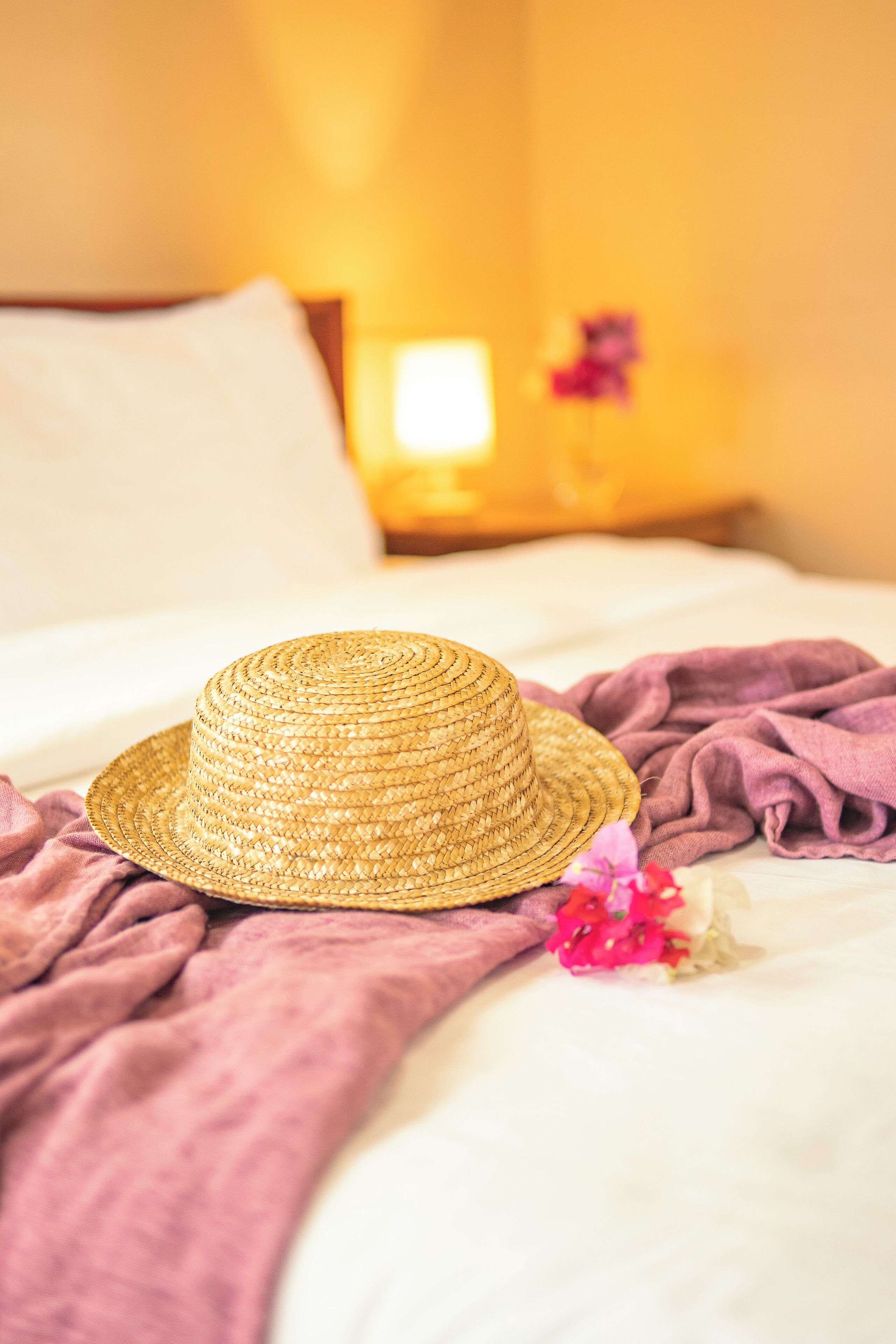 A straw hat on a bed with a purple blanket and a pink flower.