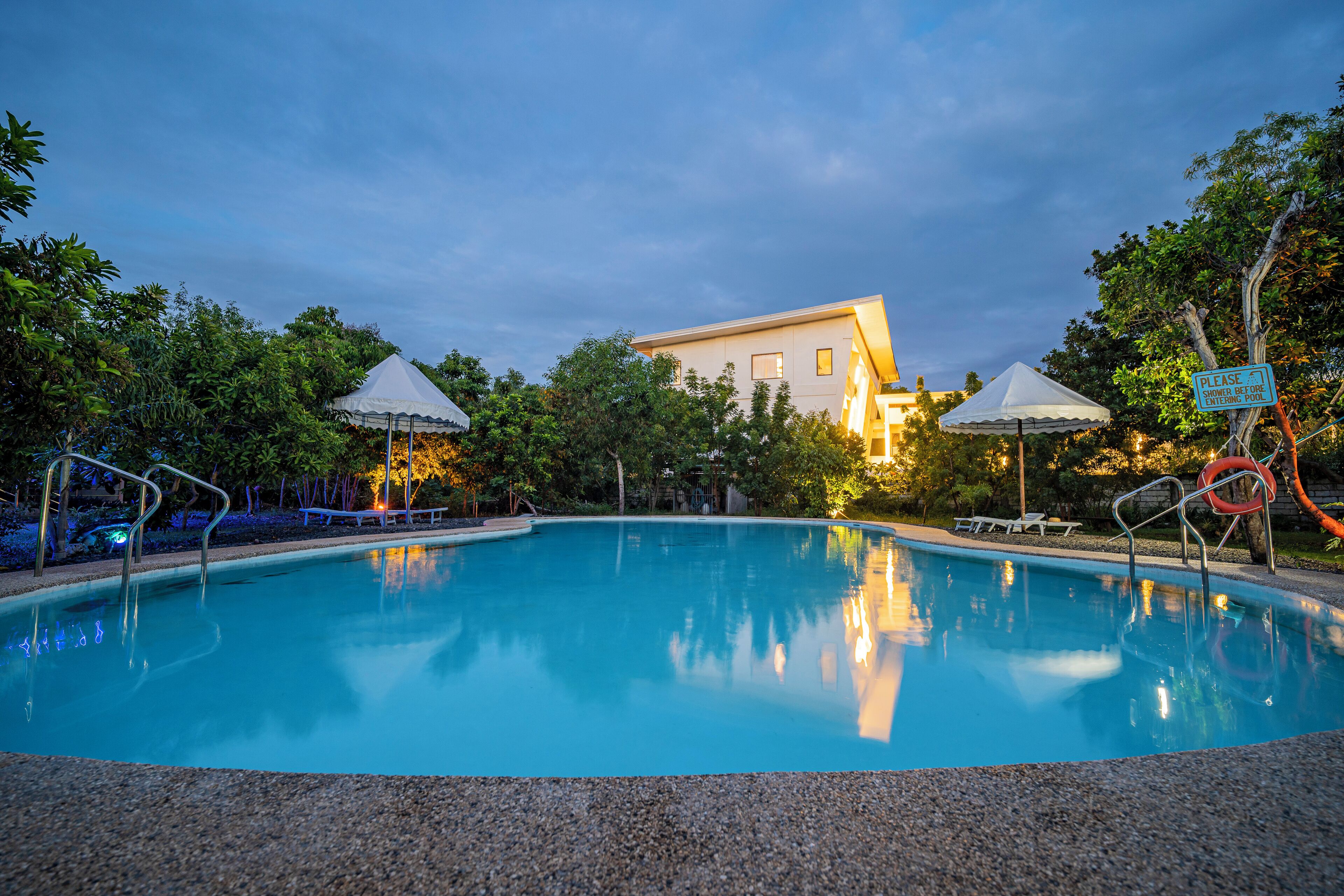 Piscina con sillas de descanso y sombrillas, rodeada de árboles y un edificio al fondo.
