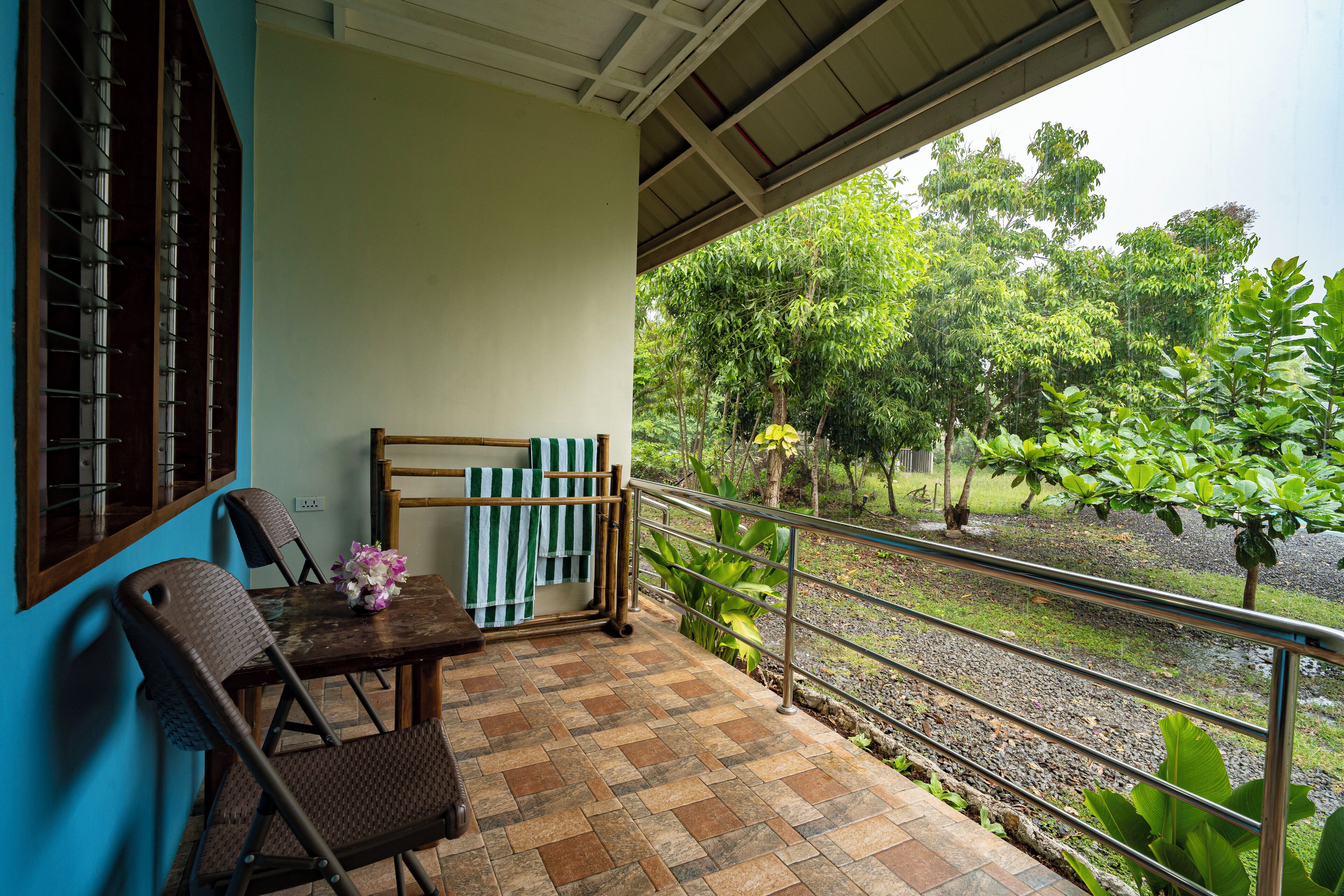A balcony with a tiled floor, a table with chairs, a striped towel, and a view of a garden with trees and a water body.