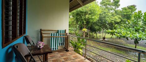 A balcony with a tiled floor, a table with chairs, a striped towel, and a view of a garden with trees and a water body.