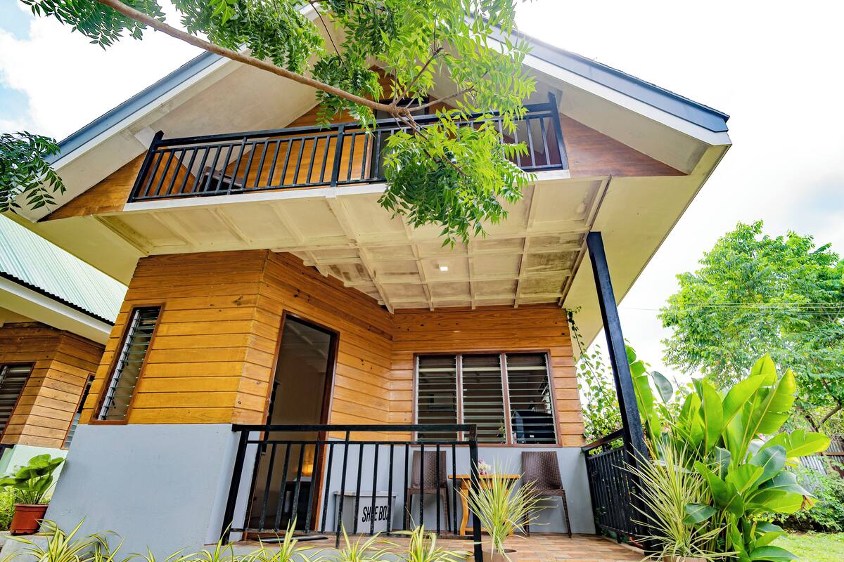 A two-story wooden house with a balcony, a covered porch, and potted plants.