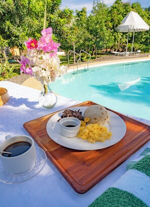 A dining table with a plate of food, a cup of coffee, and a vase of flowers by a pool. - Ana-as Apartelle  (Panglao)