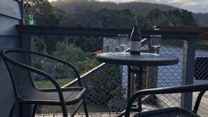 A wooden deck with two wicker chairs and a small round table, overlooking a body of water and a forested hillside. - A cosy garden cottage, just 400m to the surf beach. (Bicheno)