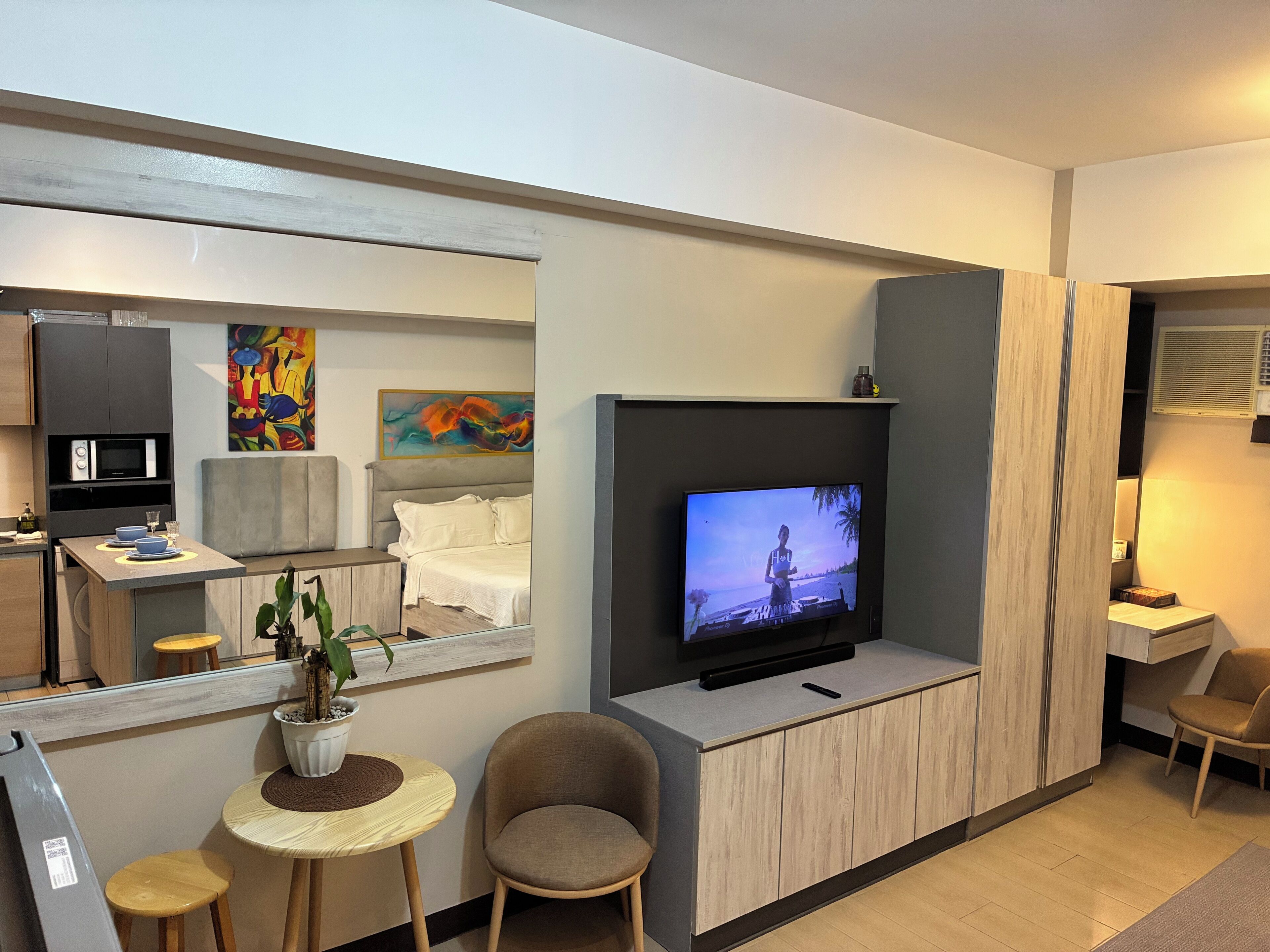 A modern hotel room with a large flat-screen TV, a dining area with a microwave, a small table with a plant, and a seating area with a chair.