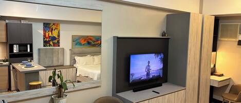 A modern hotel room with a large flat-screen TV, a dining area with a microwave, a small table with a plant, and a seating area with a chair.