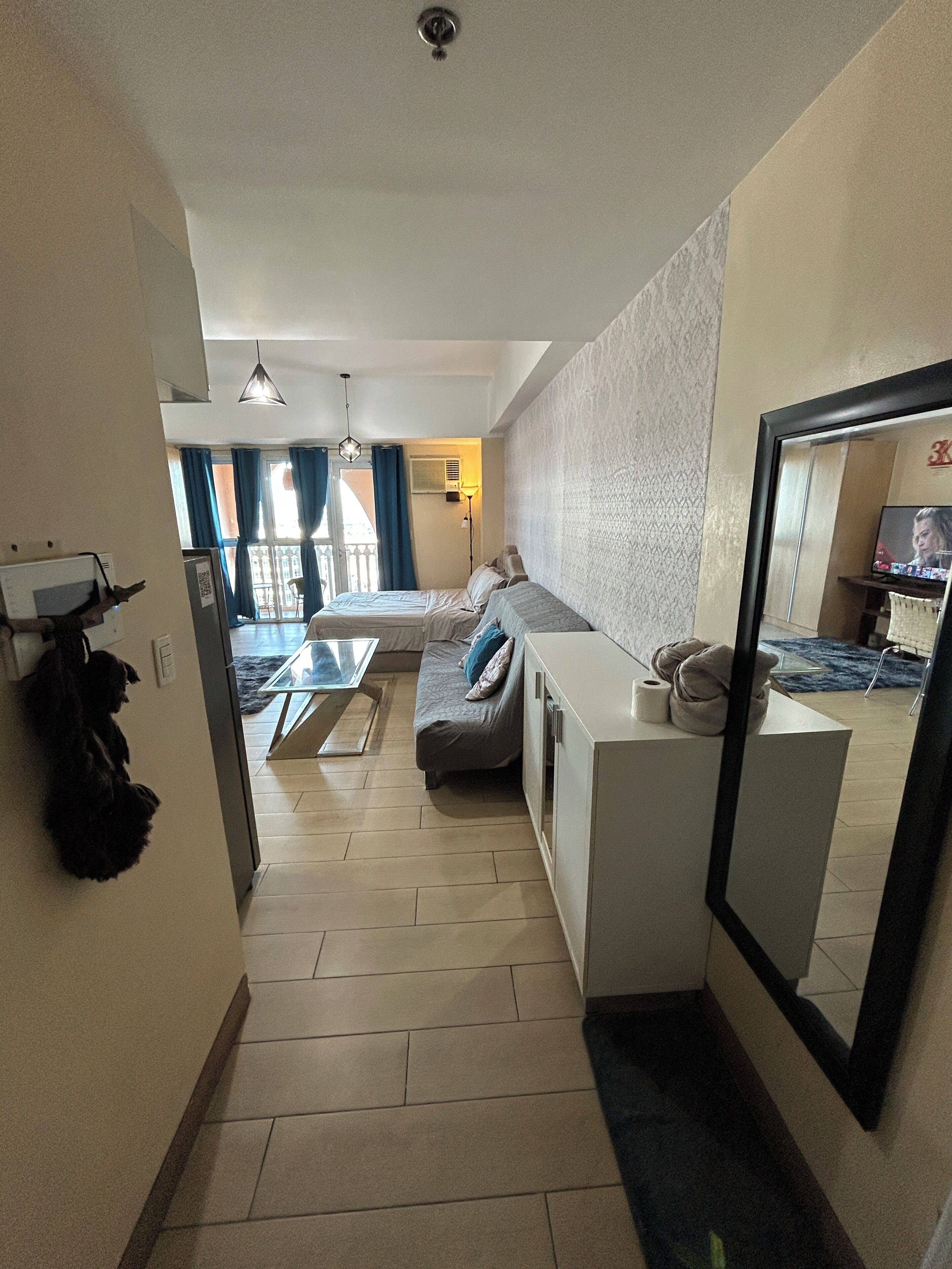 Une chambre d’hôtel compacte comprenant un coin cuisine, un lit avec une couette grise, un petit canapé et un grand miroir.