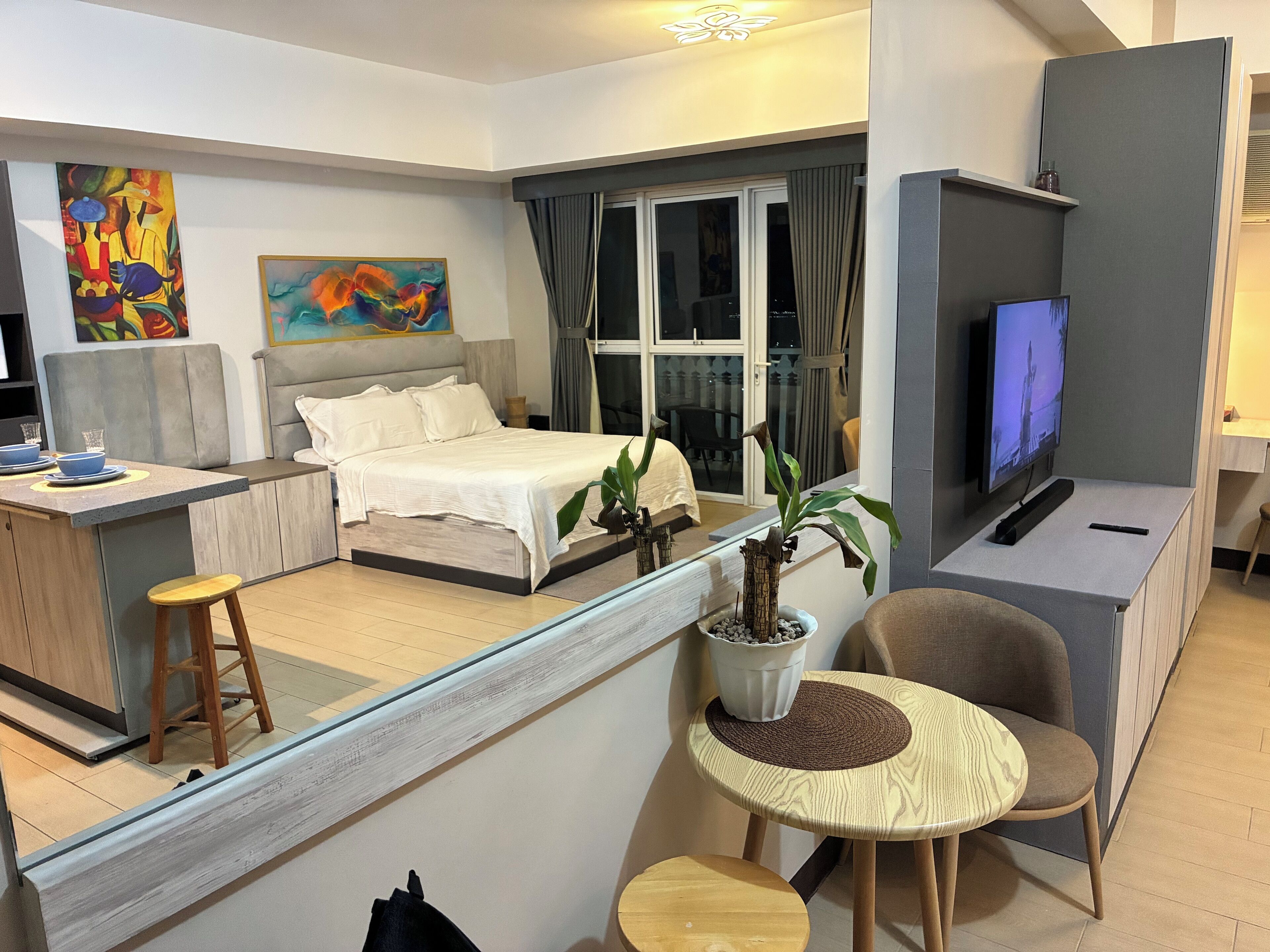 A modern hotel room with a bed, a TV, a dining area, and a balcony with a view.