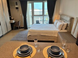 Habitación de hotel con cama, escritorio, silla, mesa de comedor con platos y vasos, y vistas a edificios a través de la ventana.