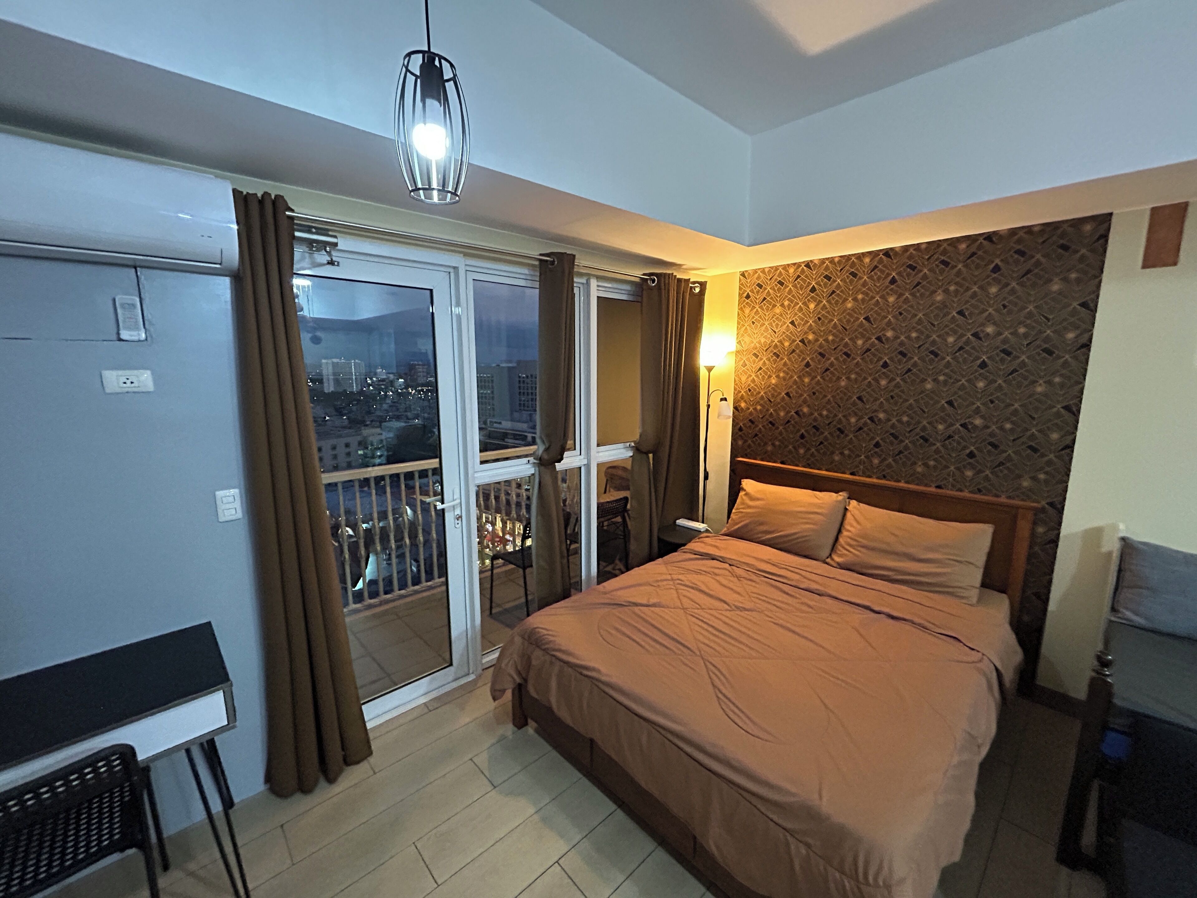 Une chambre avec un lit, un bureau, une chaise, un balcon avec des rideaux et une vue sur la ville.