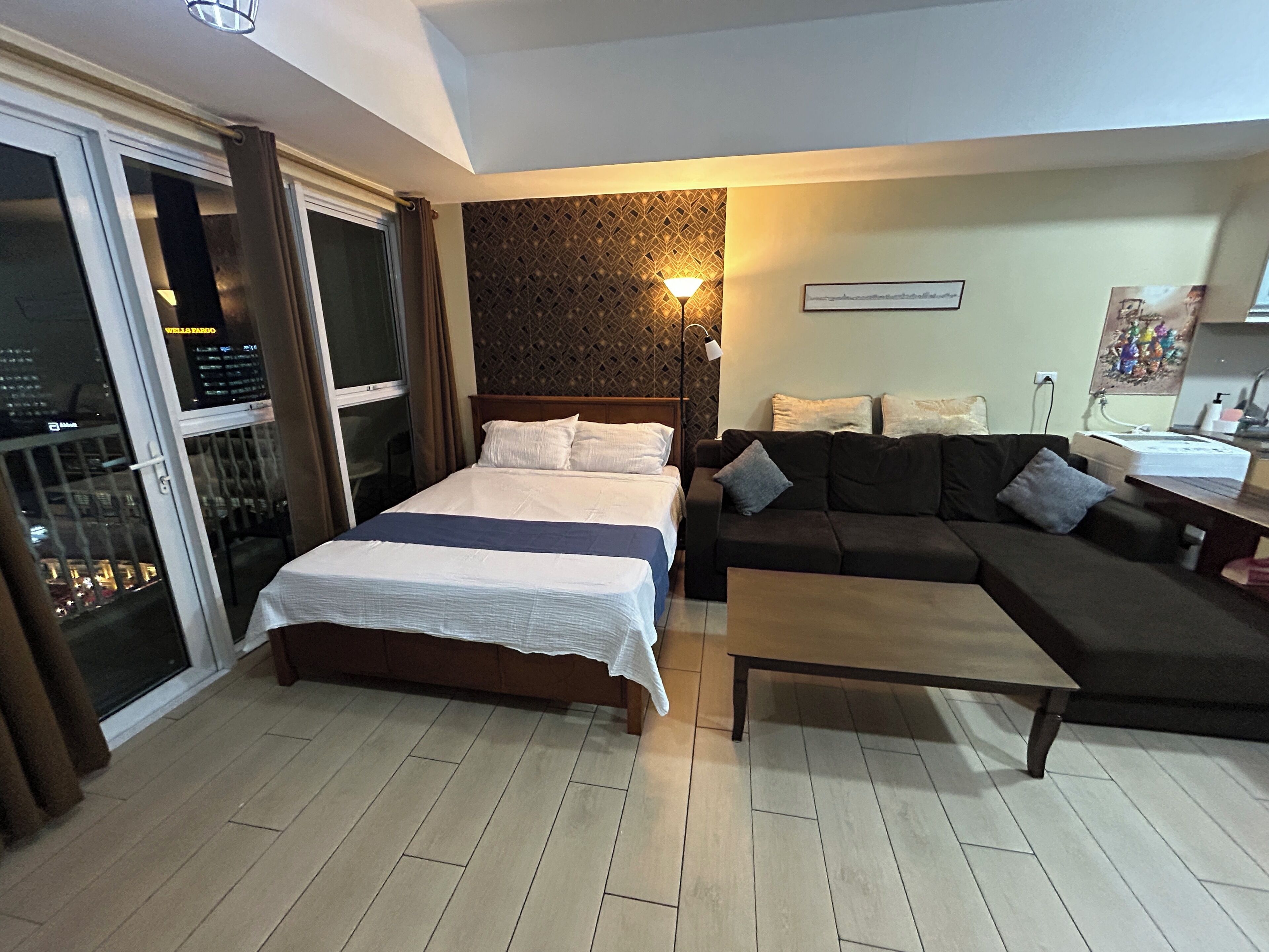 A hotel room with a bed, a sofa, a coffee table, a wall-mounted lamp, and a view of the city through a glass door.