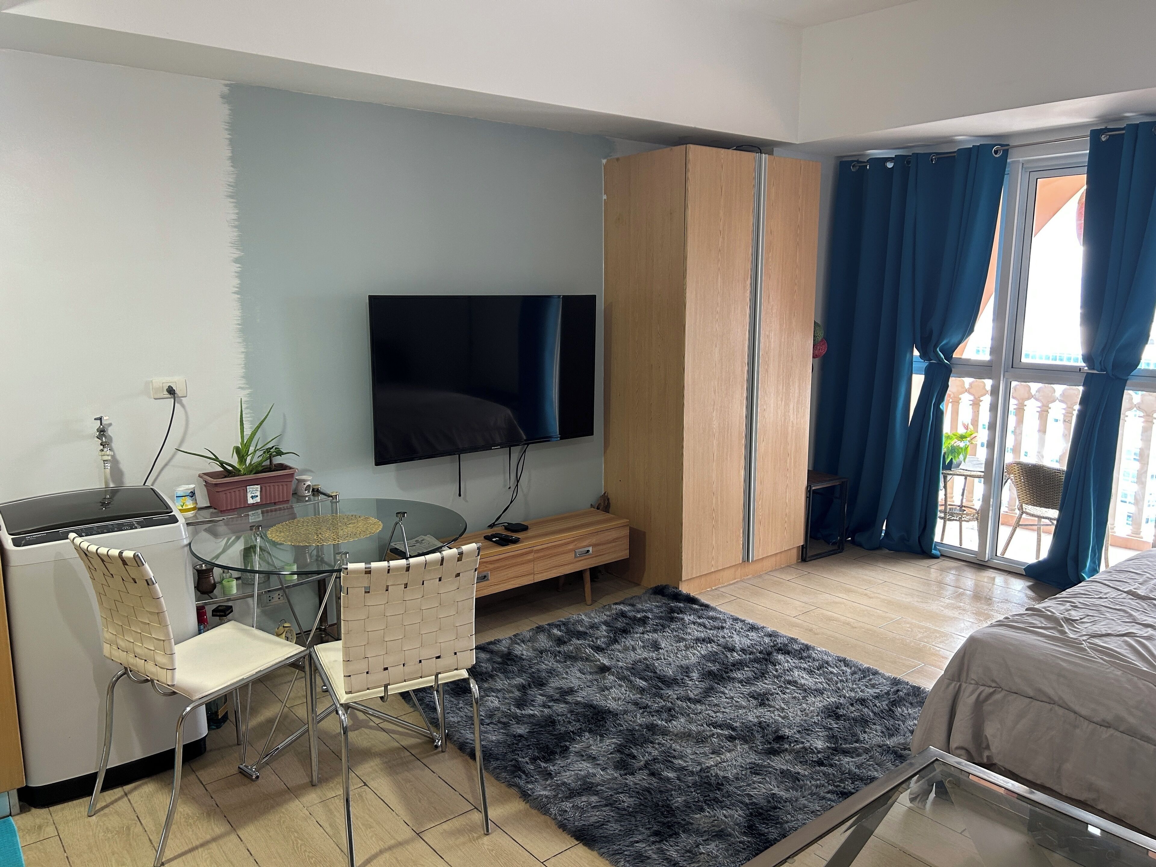 A modern bedroom with a bed, a TV, a wardrobe, a glass table with chairs, and a balcony with blue curtains.