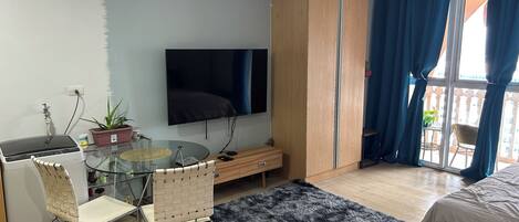A modern bedroom with a bed, a TV, a wardrobe, a glass table with chairs, and a balcony with blue curtains.