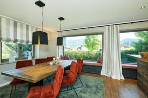 A dining area with a wooden table, red chairs, pendant lights, and a view of greenery and buildings through large windows.