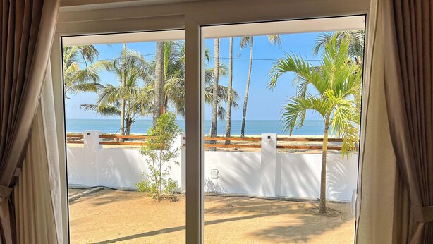 A room with a view of a beach and palm trees through a window with curtains. - Private Beach Front Villa (Thalikulam)