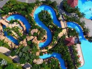 Une grande piscine sinueuse, bordée d’une végétation luxuriante et de rochers, avec un bâtiment aux toits rouges situé à proximité.