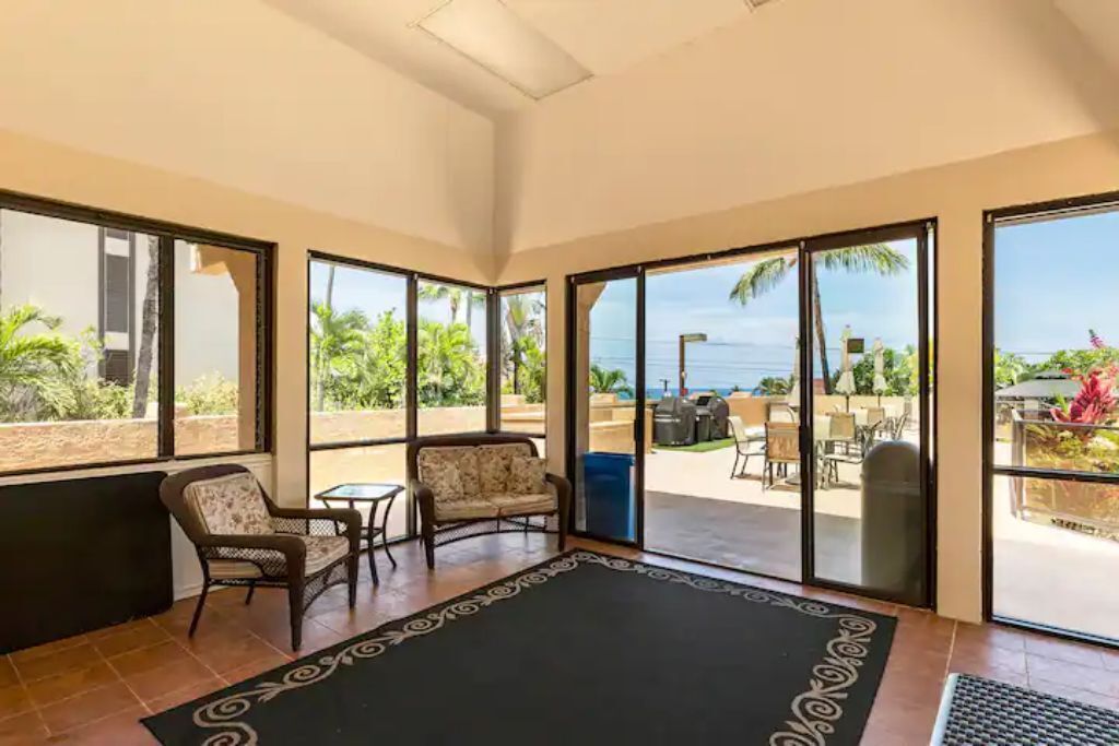 Una habitación con un gran ventanal corredizo que da a un patio exterior con vistas al océano. Hay dos sillones de mimbre y una mesita en la habitación.