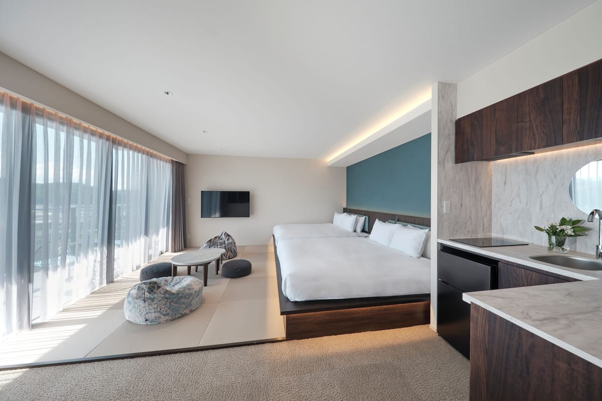 Habitación de hotel moderna con una cama grande, una pequeña zona de estar, un área de cocina y un amplio ventanal con cortinas.
